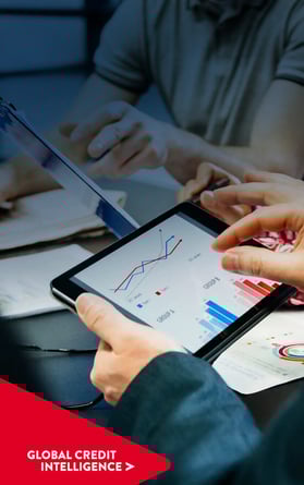 Team of business people reviewing analytics on a tablet and chart. Global Credit Intelligence chevron point on bottom left corner overlaid.