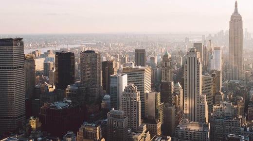 An aerial view of New York city skyline at sunset, featuring numerous skyscrapers and high-rise buildings. The Empire State Building is visible on the right, bathed in warm golden light, with a hazy urban landscape extending into the distance.