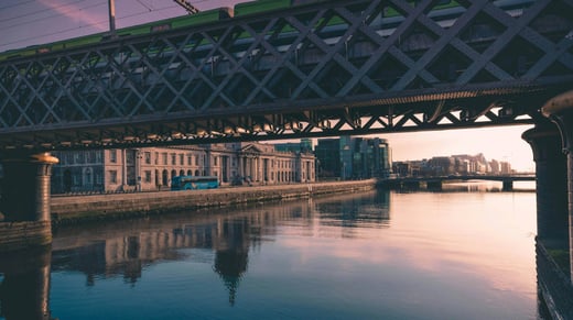A scenic view of the River Liffey in Dublin, Ireland, at sunset, featuring the historic Loopline Bridge with a train passing overhead. Below, the calm water reflects the surrounding structures. The neoclassical Custom House building lines the riverbank, with modern glass buildings in the background. A bus is seen on the street, adding to the urban atmosphere.
