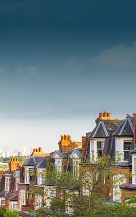A scenic view of red-brick Victorian houses in Highgate, London, with their distinctive chimneys and dormer windows. The lush greenery in the foreground contrasts with the modern skyline of Canary Wharf visible in the hazy distance.